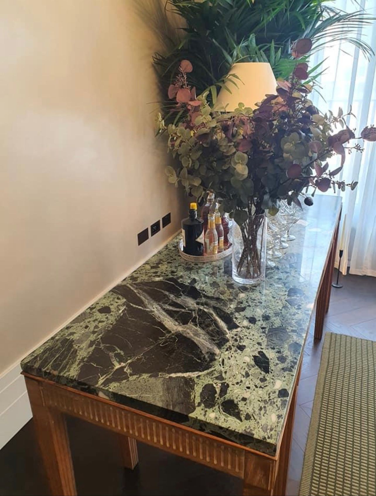 Green marble tabletop with bottles, a vase of plants, and a lamp in a bright room.