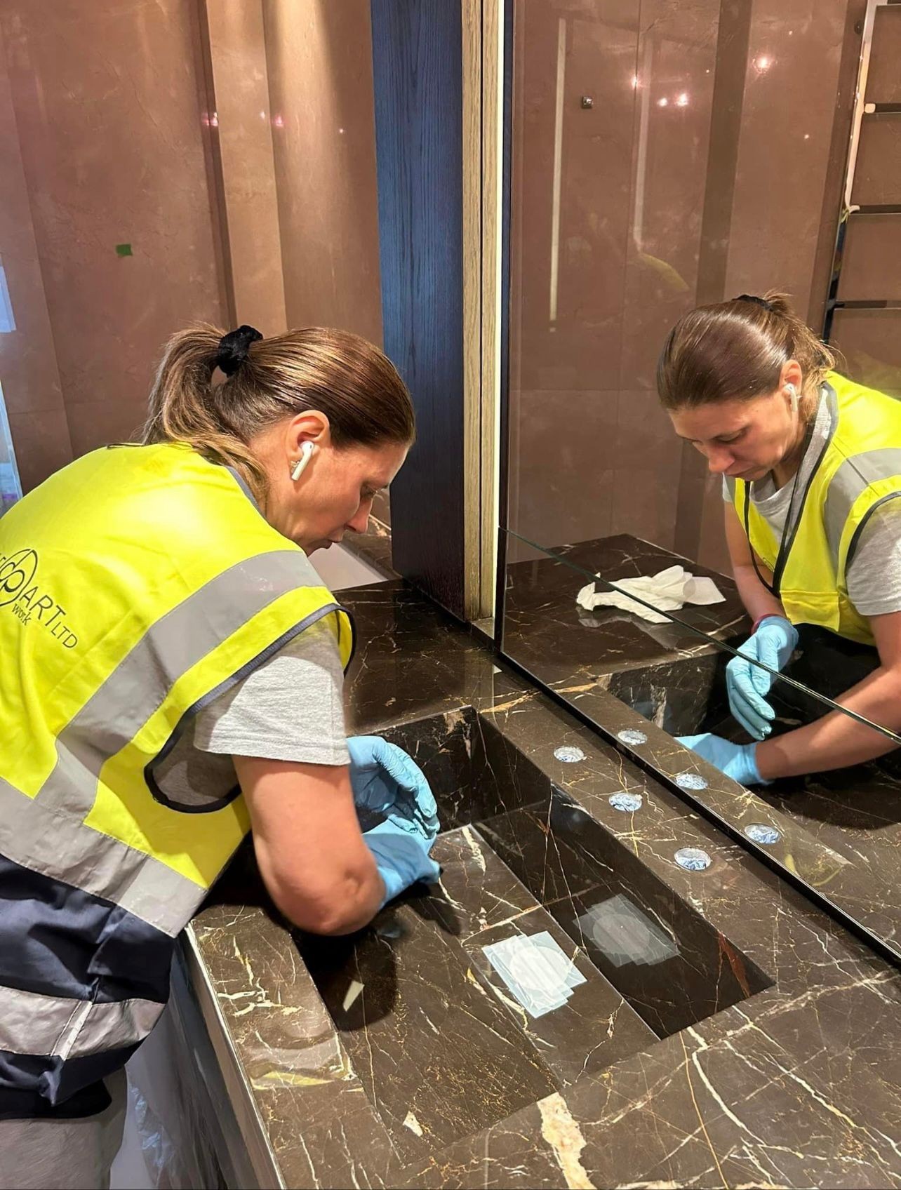 Person wearing a high-visibility vest and gloves, cleaning a marble sink in a restroom.